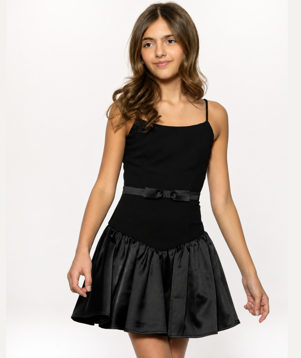 Model wearing the KatieJ NYC Tween Noreen black mini dress with fitted bodice, satin bow waist, dropped waistline, and flared skater skirt.