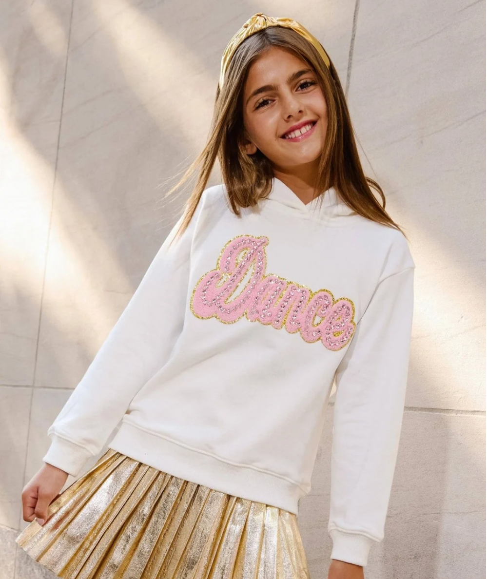 A young girl smiling while wearing a white hoodie with a pink and gold sequined 'Dance' graphic by Lola & the Boys. Tween Crystal Dance Hoodie in ivory with pink crystal Dance lettering