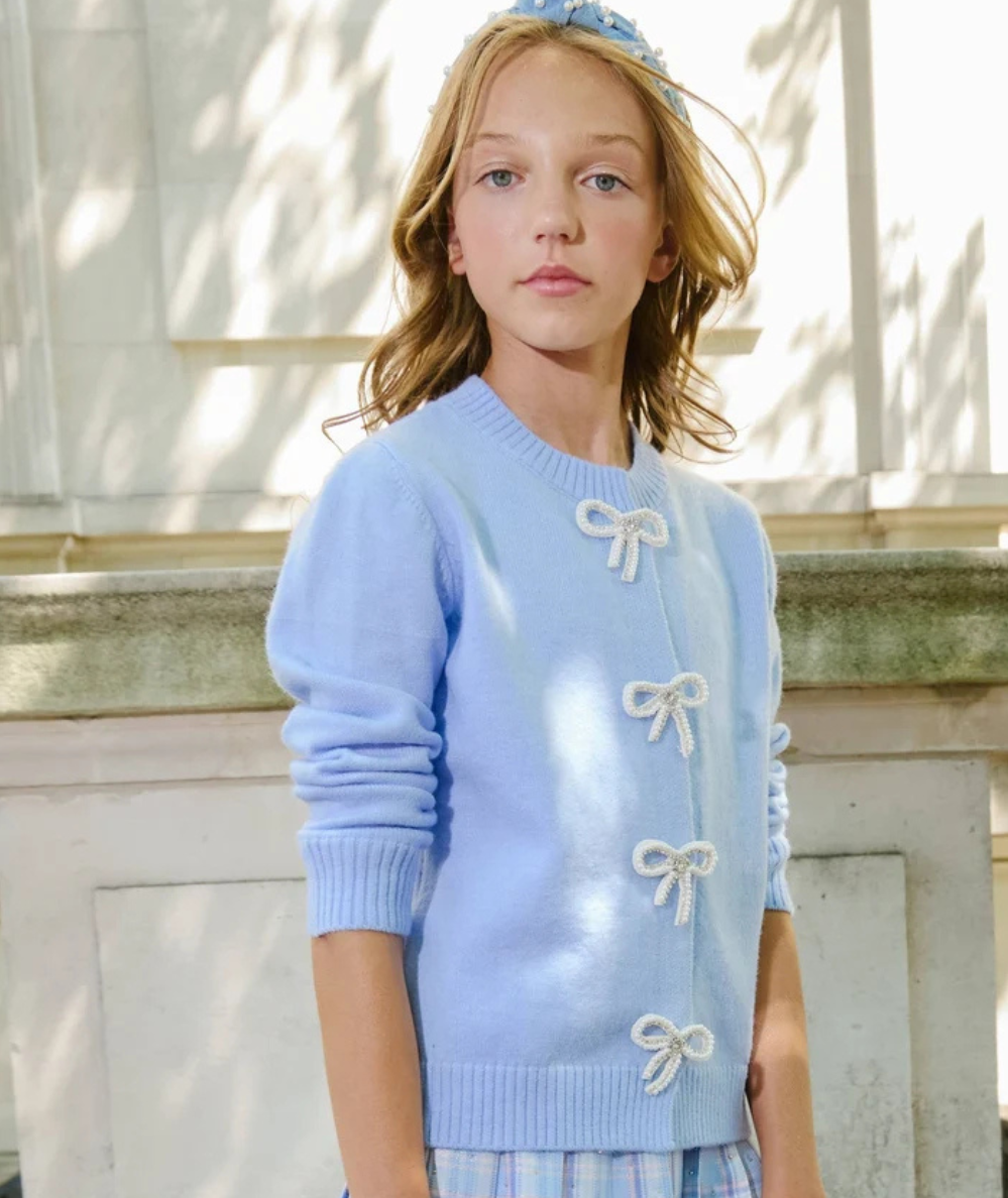 Model wearing the Lola & the Boys Tween Azure Crystal Bow Cardigan in light blue azure with four crystal bow closures and ribbed cuffs and hem.