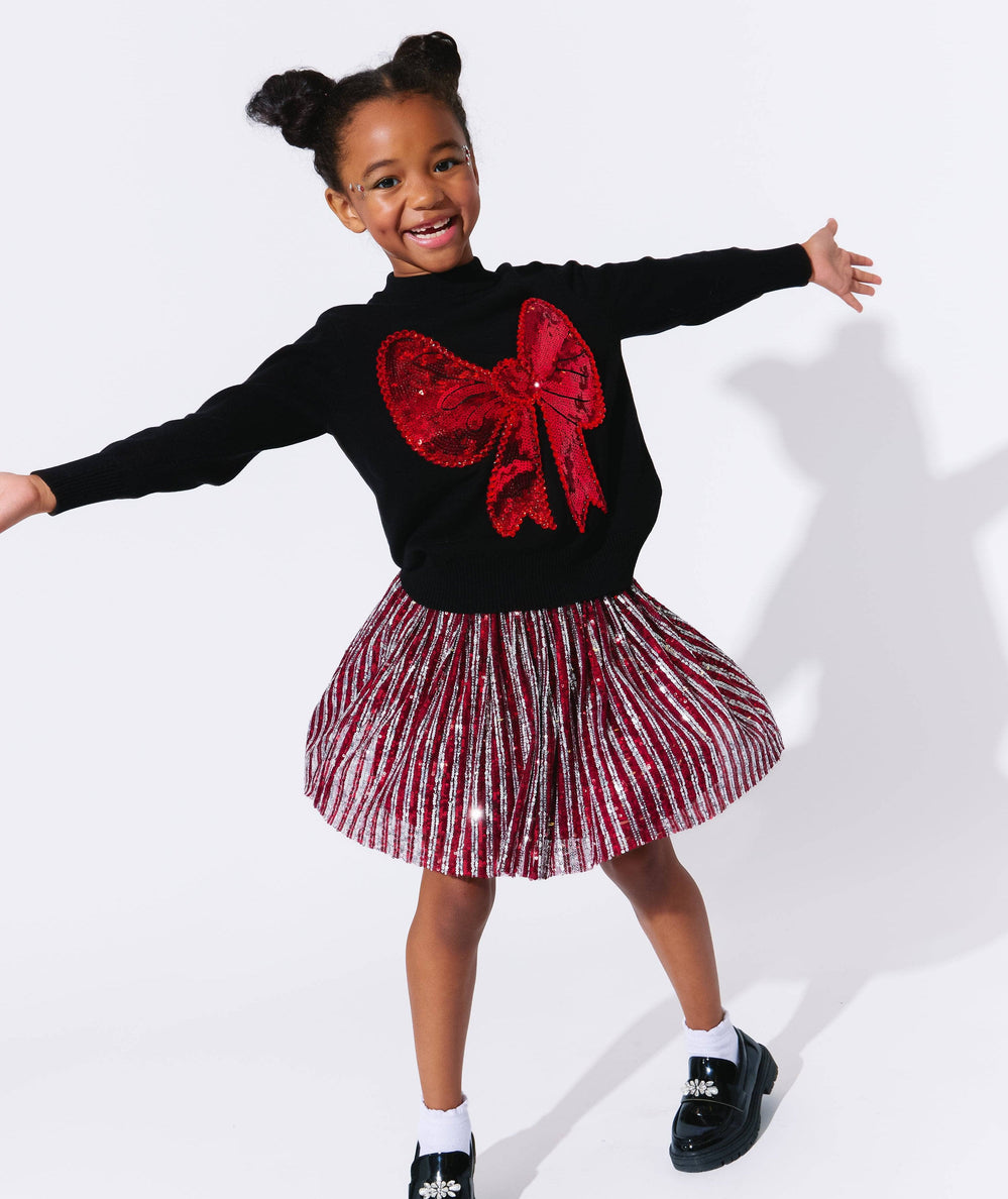 Smiling girl in the big bow sweater and a metallic pleated skirt by Lola and the Boys - CAKEWISH.