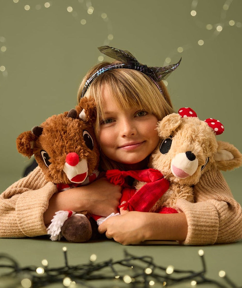 Girl hugging the Warmies Rudolph plush with holiday lights, a cozy Christmas gift - CAKEWISH.